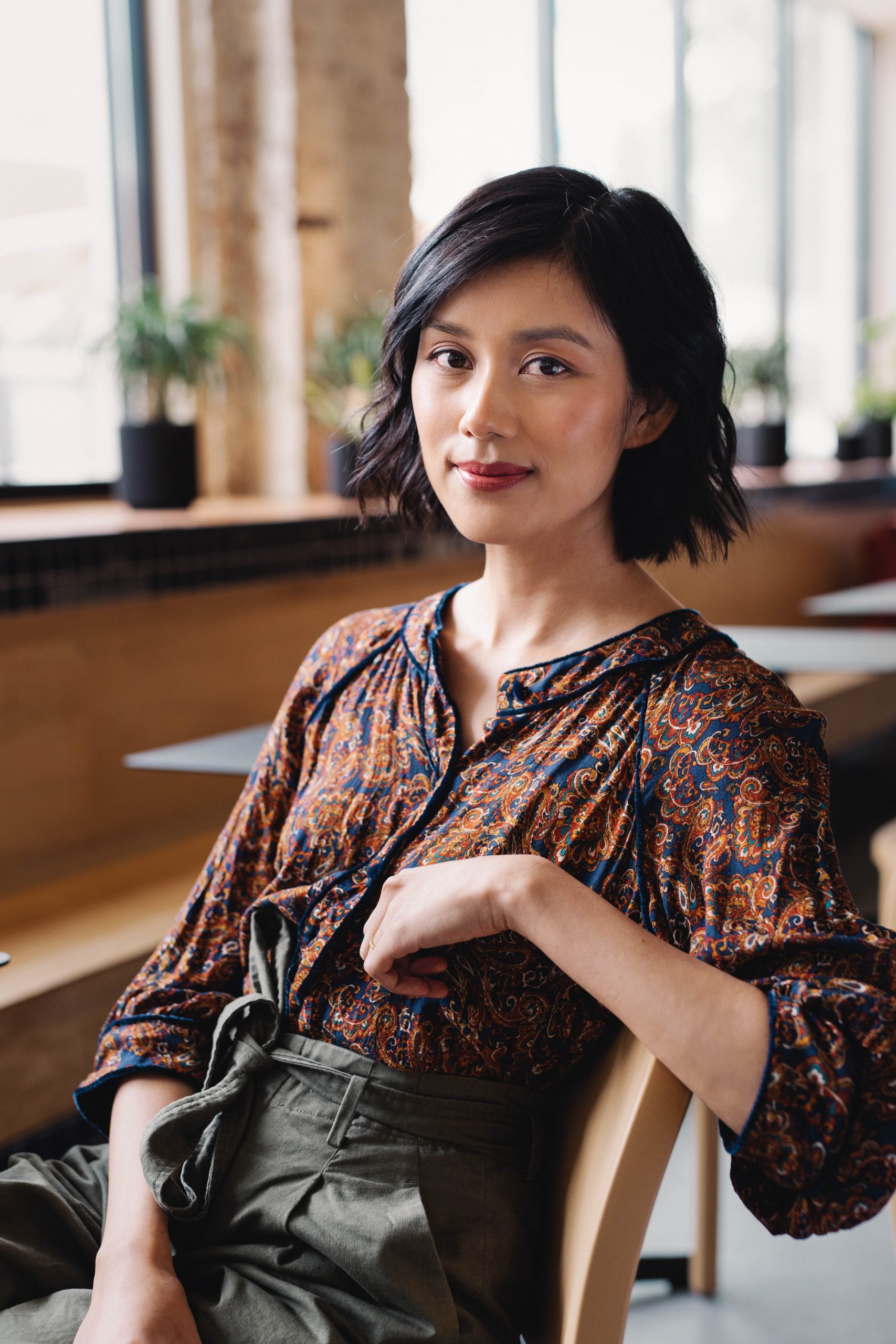 Tracey Lien sitting in a chair in a well lit space with out of focus plants in pots by windows in the background