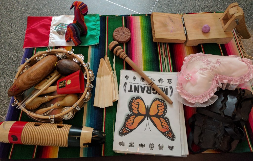 Image of a table containing a variety of objects including a Mexican flag, a pink heart pillow, and musical instruments like a cowbell and a tambourine.