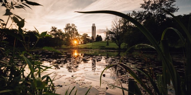 Image of Potter Lake in the foreground. The Campanile is in the background in the center, and the sun is rising to the left. 