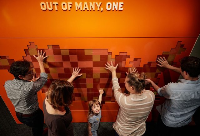 A group of people placing hands on textured blocky map of the United States with text above saying 'Out of many, one'