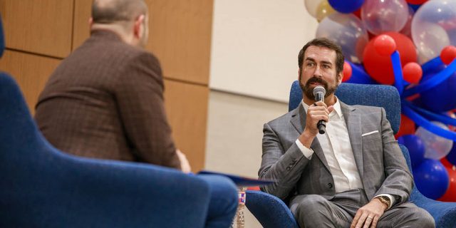 Rob Riggle on stage in a chair with a microphone