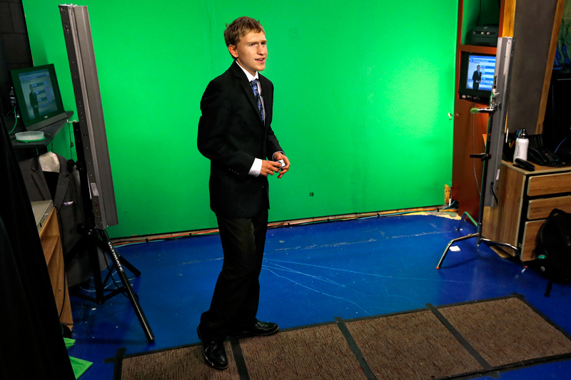 Carson, wearing a suit and tie, stands in front of a green screen